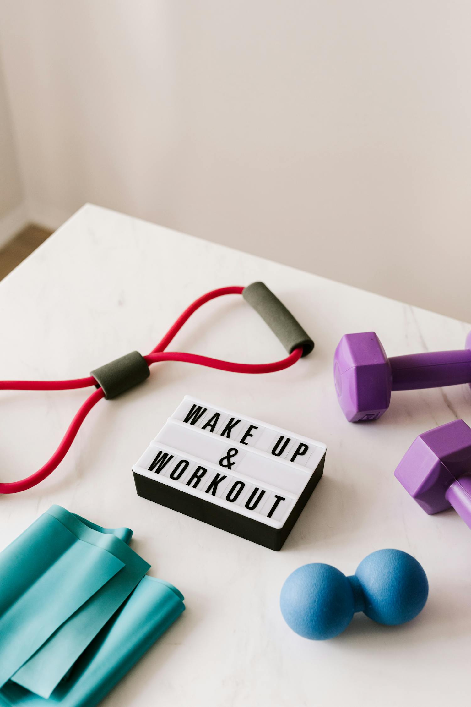 From above composition of dumbbells and massage double ball and tape and tubular expanders surrounding light box with wake up and workout words placed on white surface of table