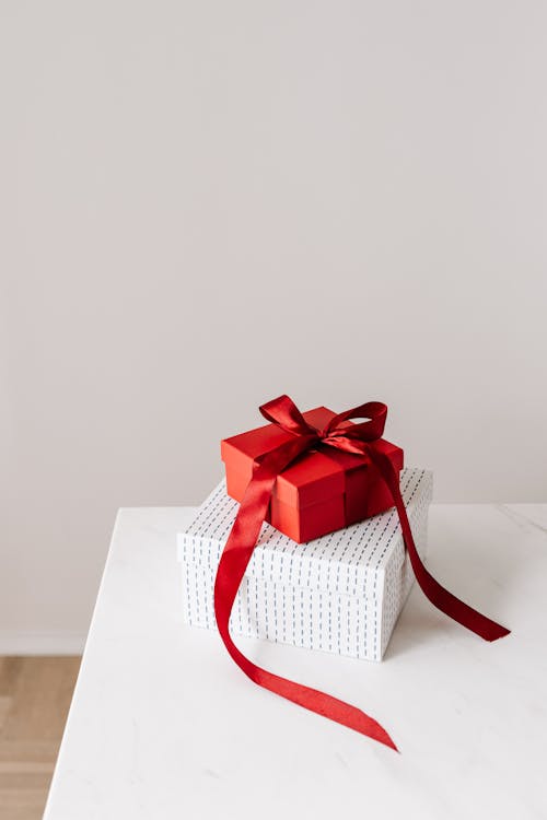 From above of wrapped with ribbon red square gift box placed on white with silver little rhombuses square box on white table against gray background