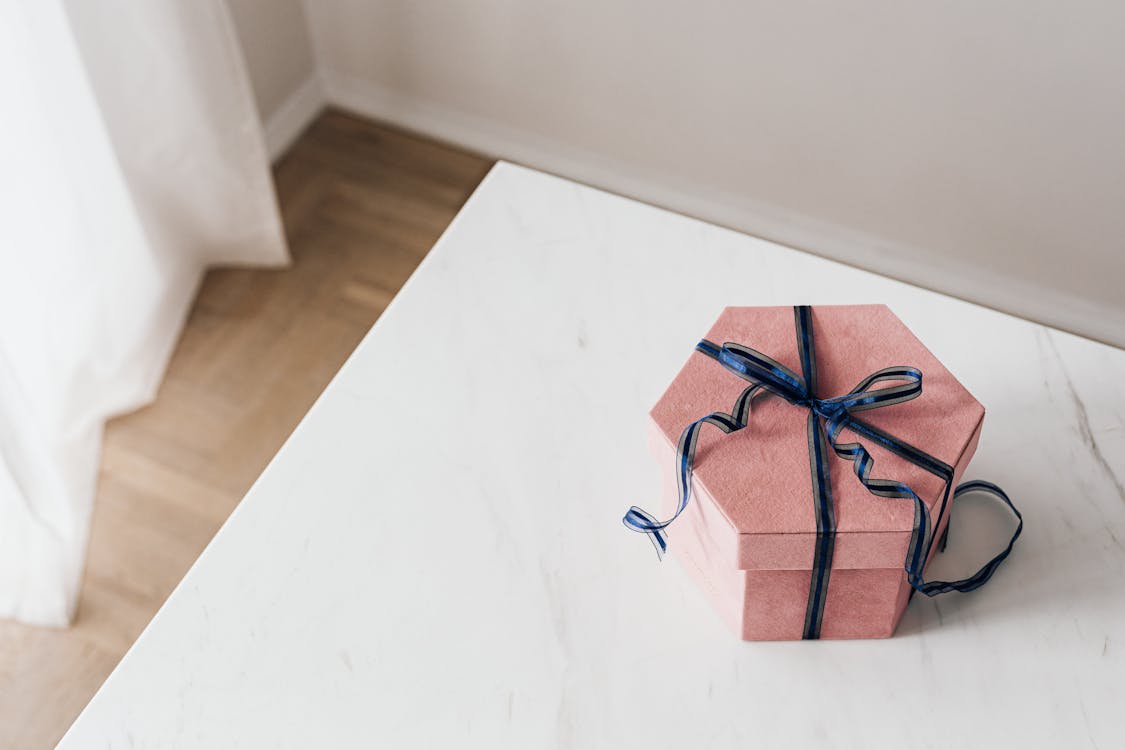 Free From above of packed pink hexagonal gift box tied with blue ribbon placed on marble table against beige wall and wooden floor and white curtain Stock Photo