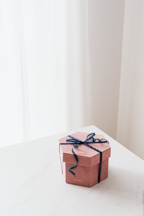 Free From above of blue ribbon tying bow on pink hexagonal gift box placed on white table against beige wall and white curtain Stock Photo