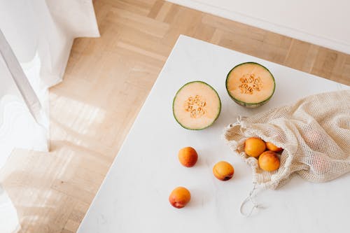 Top view of halved sweet melon and cotton sack with ripe apricots placed on white table in cozy kitchen