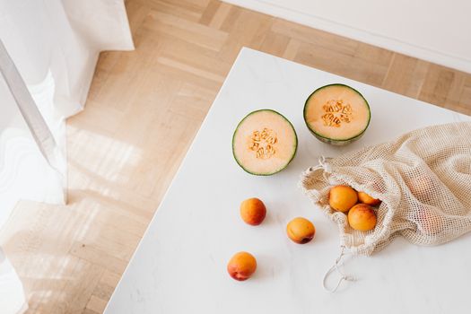 Top view of halved sweet melon and cotton sack with ripe apricots placed on white table in cozy kitchen