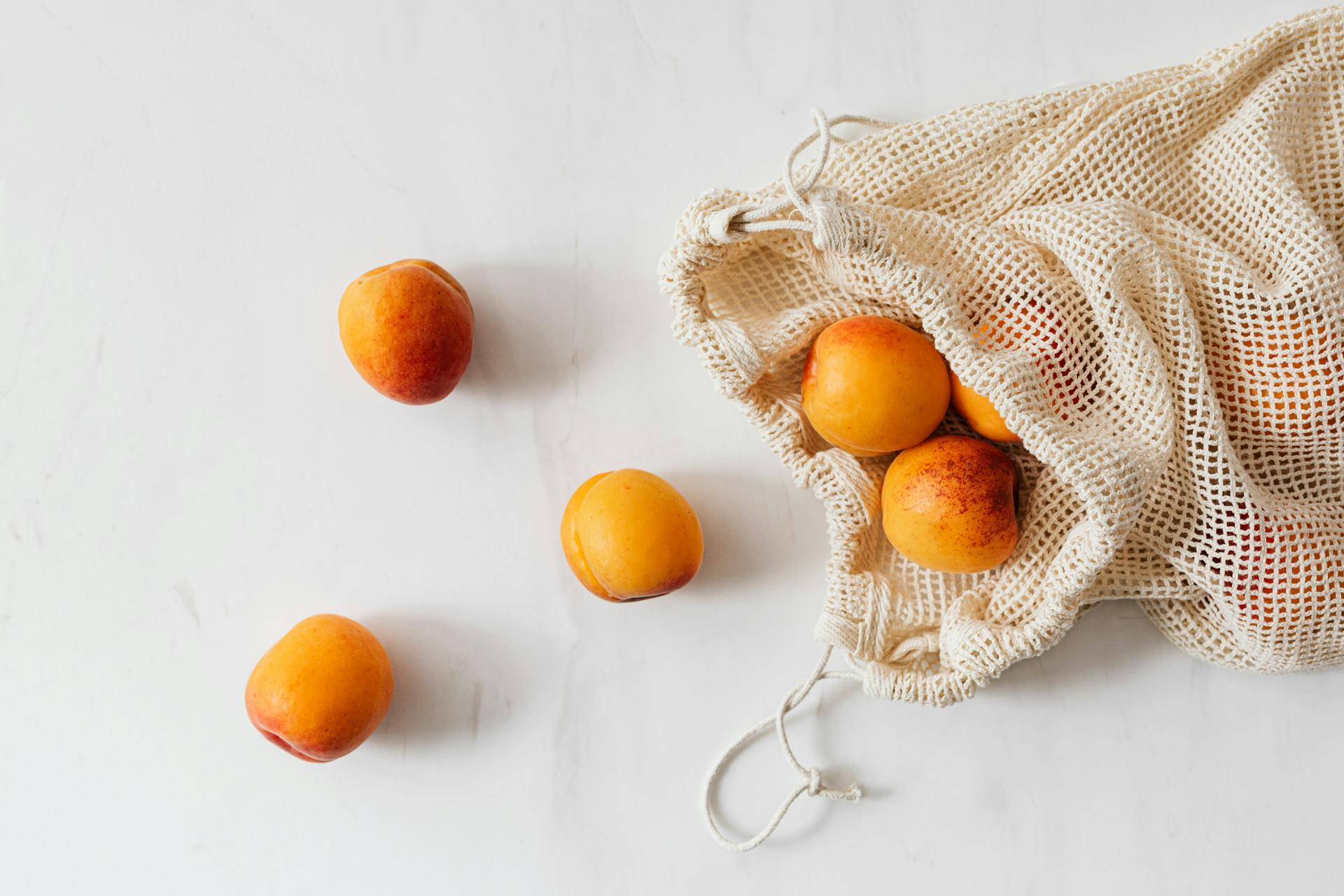 Fresh peaches spilling out of a reusable mesh bag on a white background.
