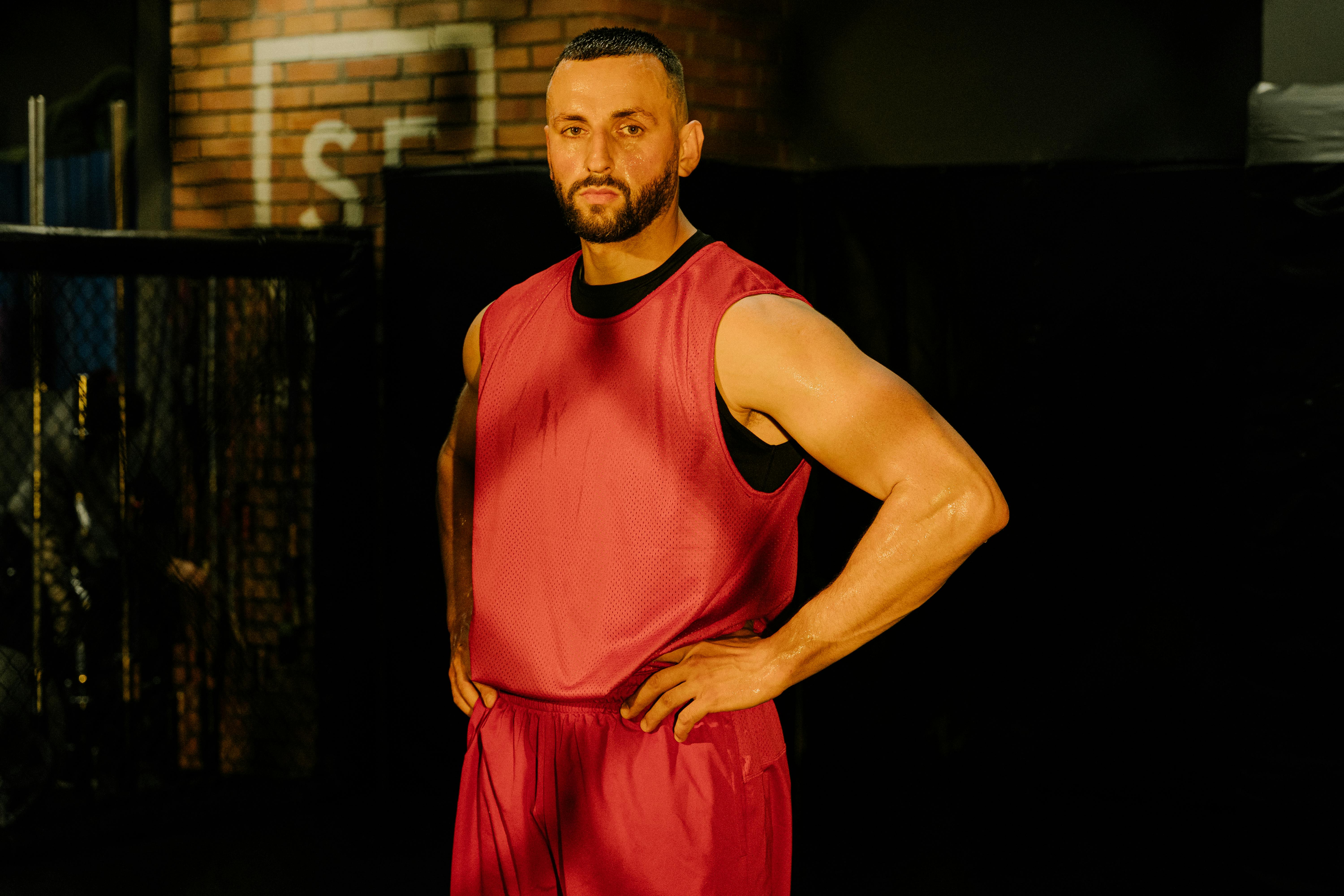 Bearded man in red activewear stands confidently with hands on waist indoors, looking determined.