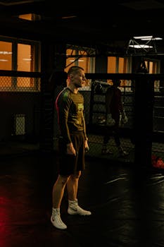 A man in activewear stands in a dimly lit boxing gym, ready for training.