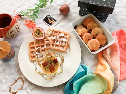 Top view of composition with plate of baked waffles with jam and exotic fruit placed near cookies and colorful neckerchief and mugs