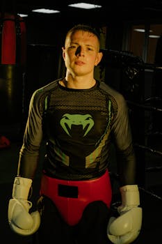 A confident male boxer wearing gloves stands ready in a dimly lit gym.