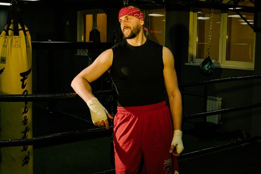 Bearded man in boxing gym with hand wraps, wearing red shorts and black tank top, posing confidently.