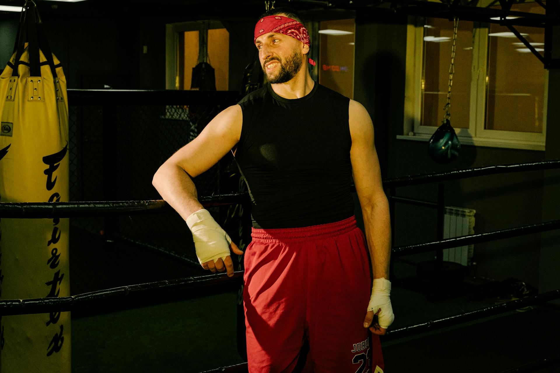 A Man in Black Tank Top and Red Pants Leaning on the Boxing Ring