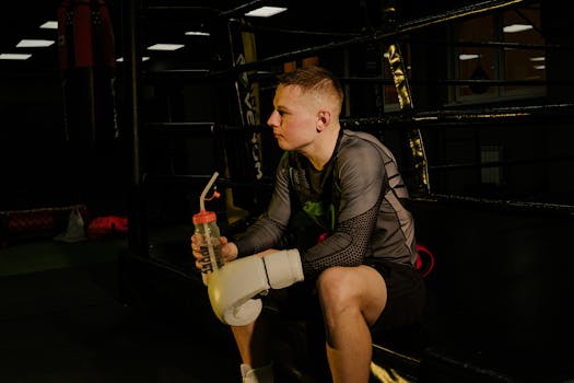 Male boxer sitting in a boxing ring, wearing gloves, and holding a water bottle.