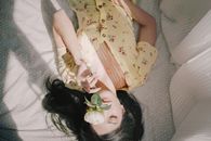 晨光中，一位穿着花裙的年轻女子放松地躺在床上，手里拿着一朵玫瑰