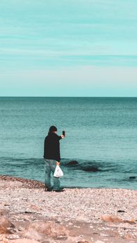 Back view of full body unrecognizable man with smartphone in hand taking photo of sea