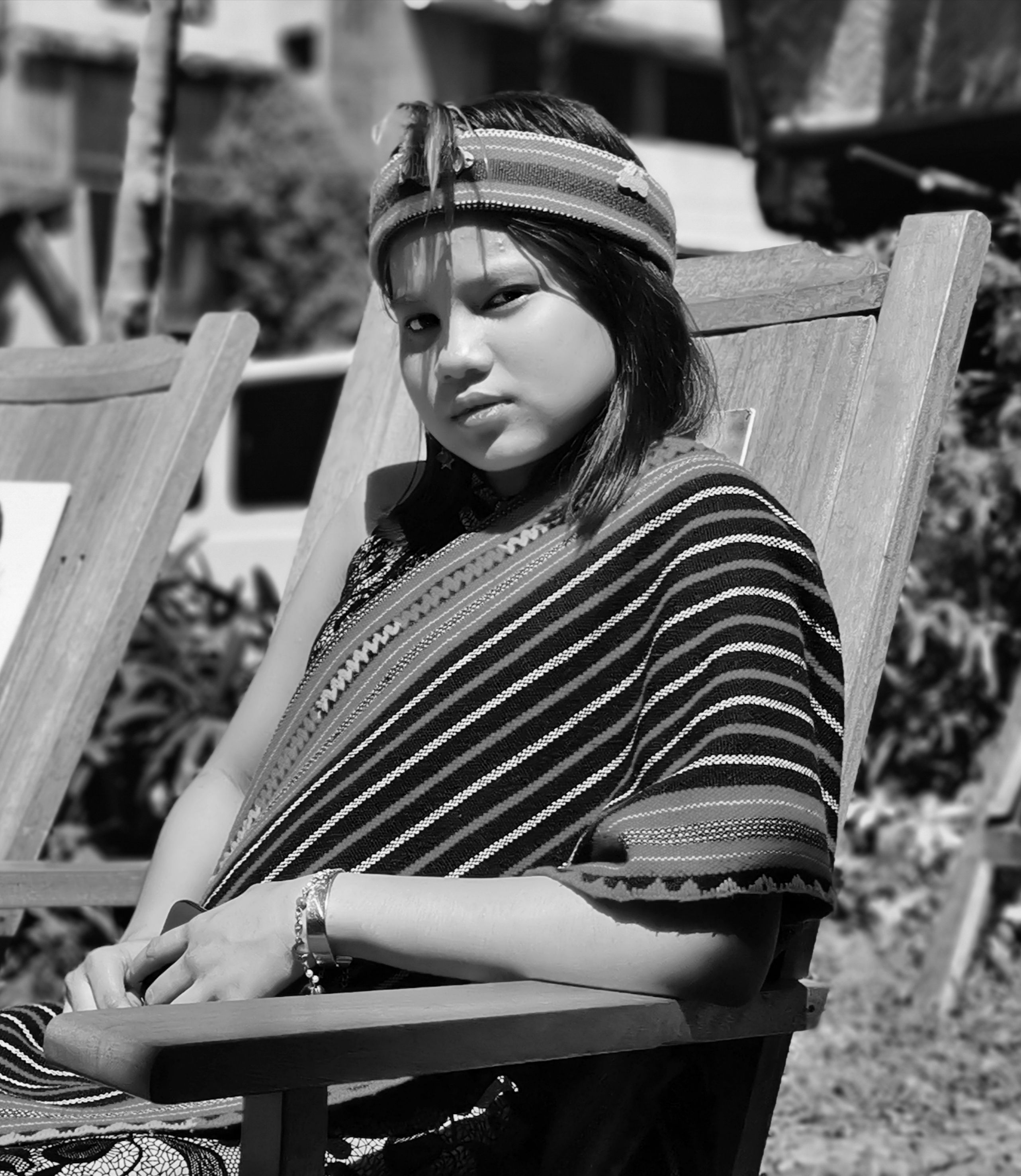 A young girl in traditional attire sitting on a wooden chair outdoors, captured in black and white.