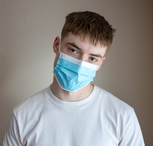Unemotional male wearing casual white shirt and protective medical mask standing against white plain wall