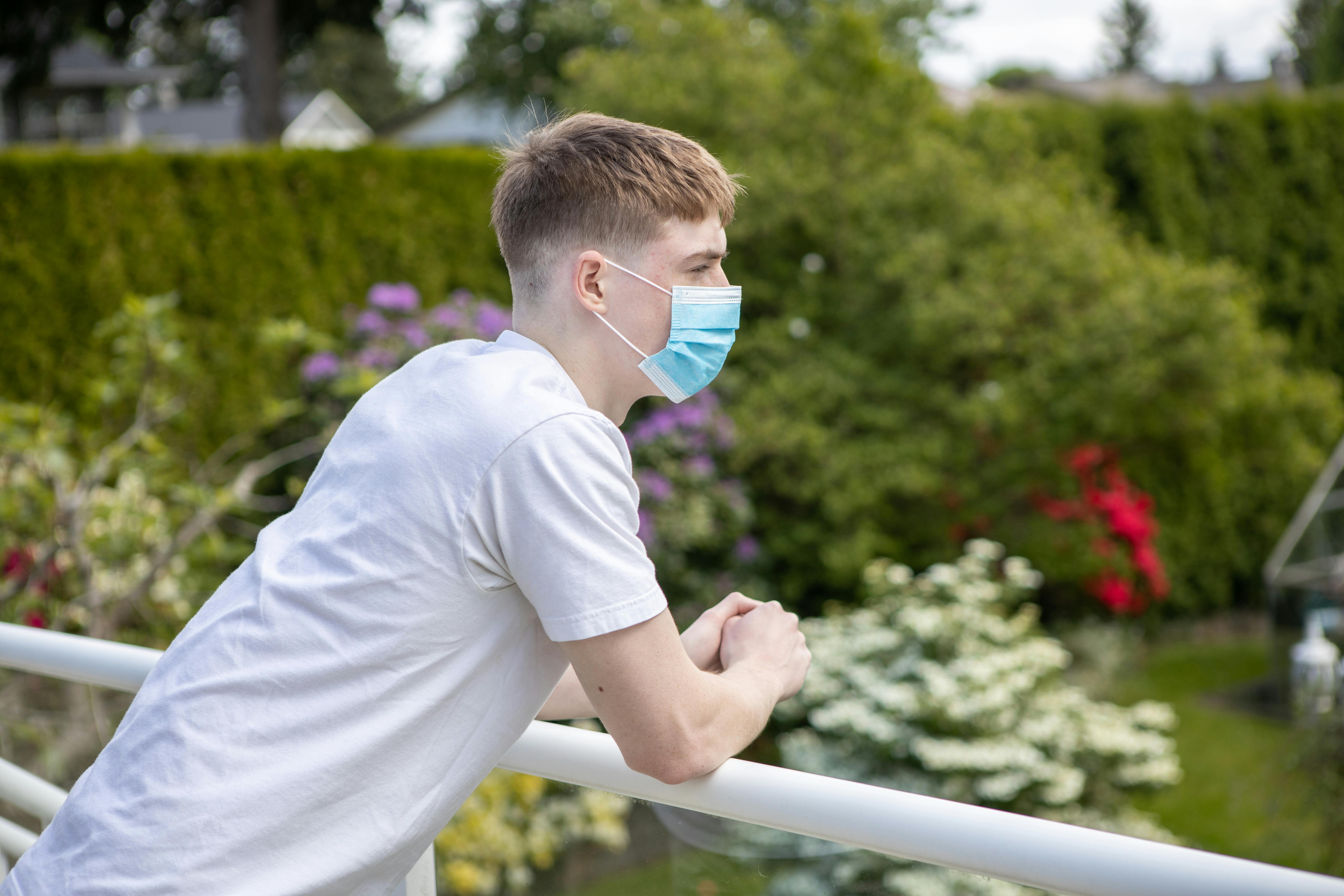 Caucasian man wearing a surgical mask and white shirt, leaning on a railing outdoors.