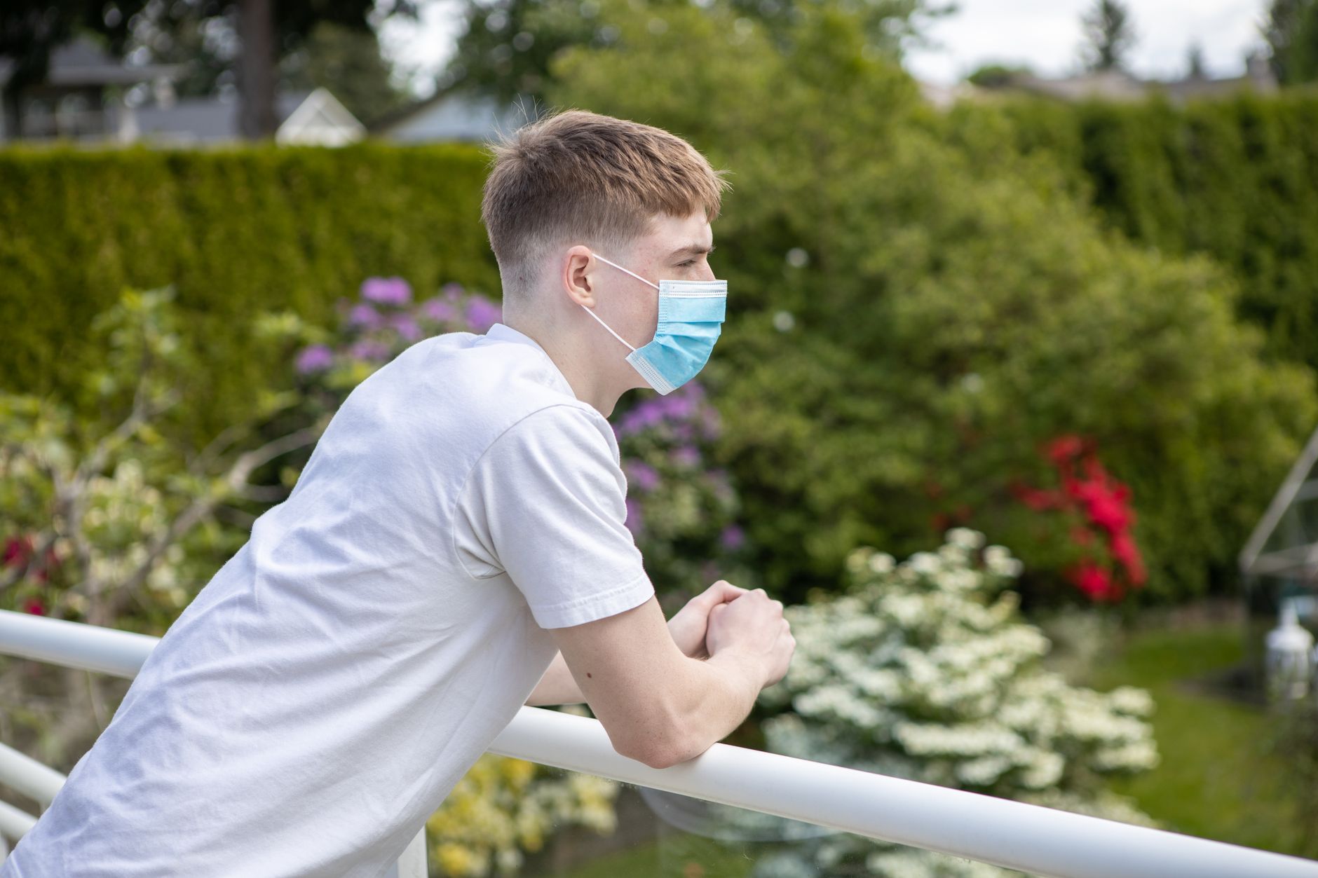 Caucasian man wearing a surgical mask and white shirt, leaning on a railing outdoors.