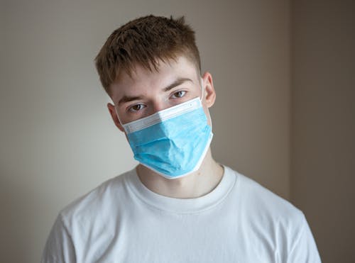 Serious young male wearing casual shirt and respirator standing against gray wall and looking at camera