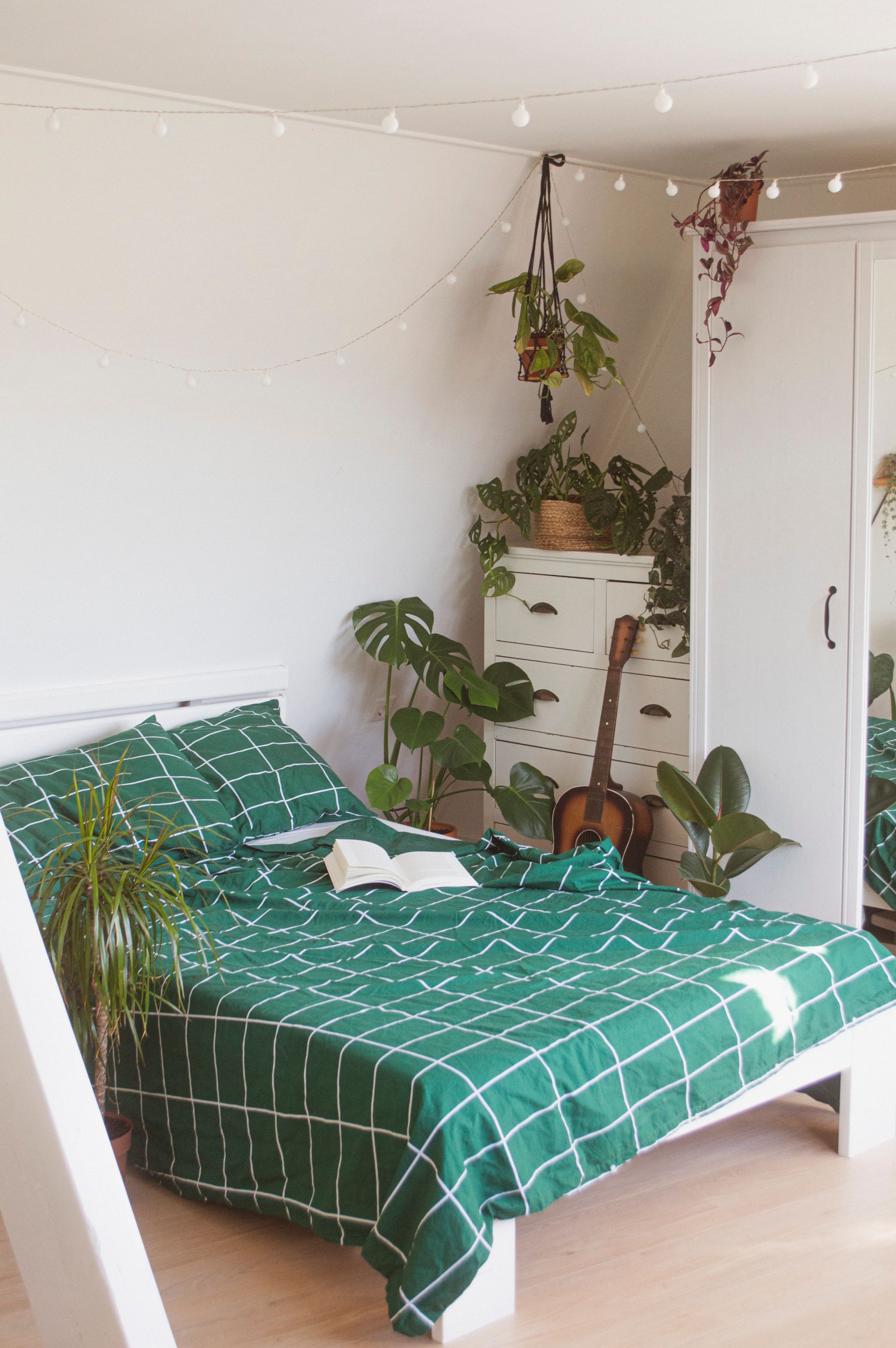 Bright bedroom with green checked bedding, a guitar, string lights and several leafy houseplants