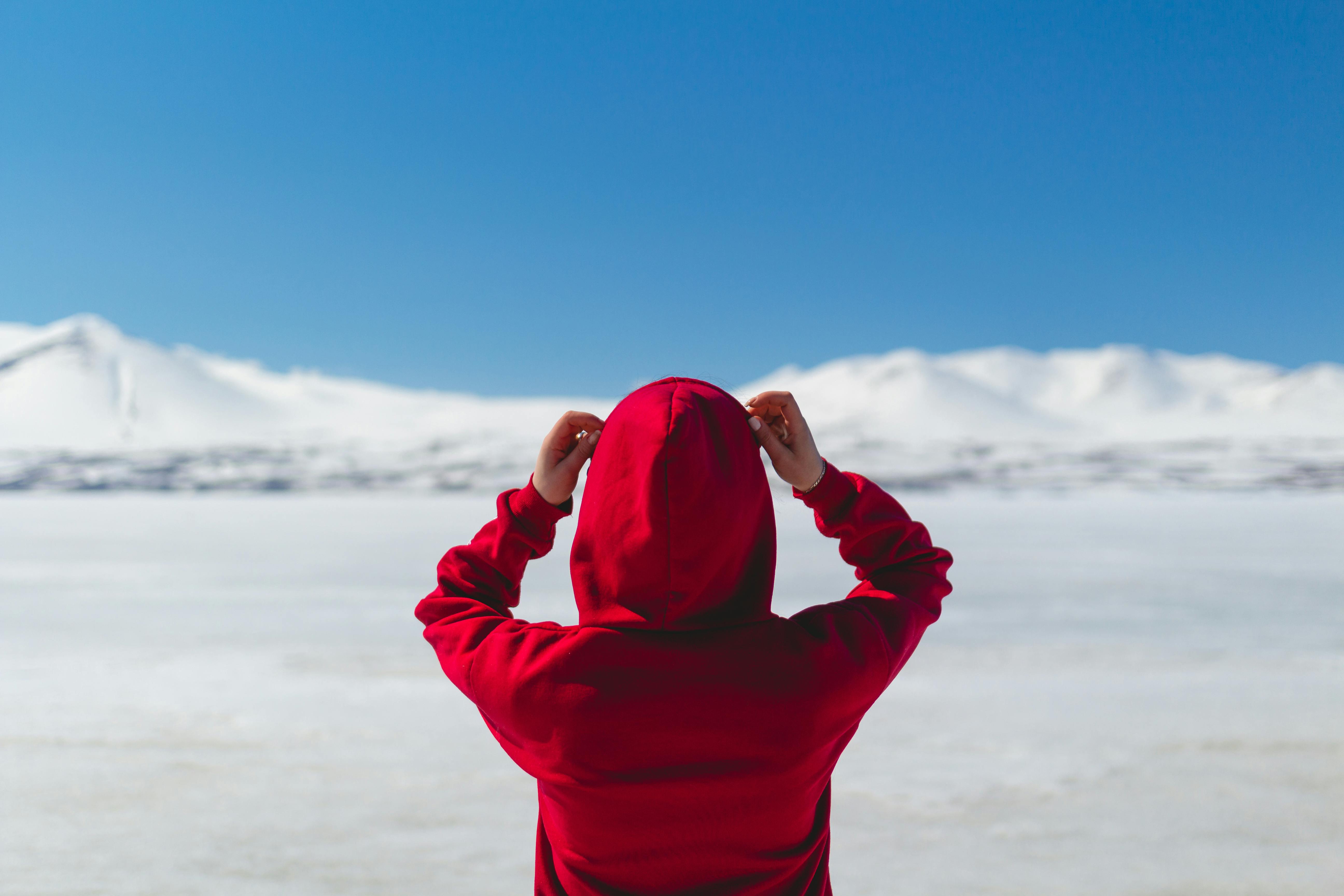 Back view of a person in a red hoodie facing snowy mountains in a remote outdoor location.