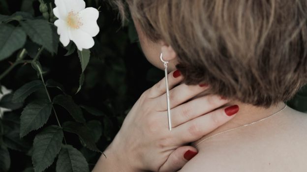 Back view anonymous female with short haircut wearing stylish earring and touching neck gently with manicured hands in summer blooming garden