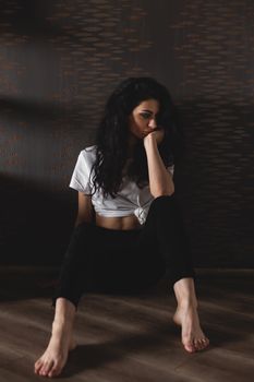 Pensive woman sitting barefoot on a wooden floor, lost in thought in a moody setting.