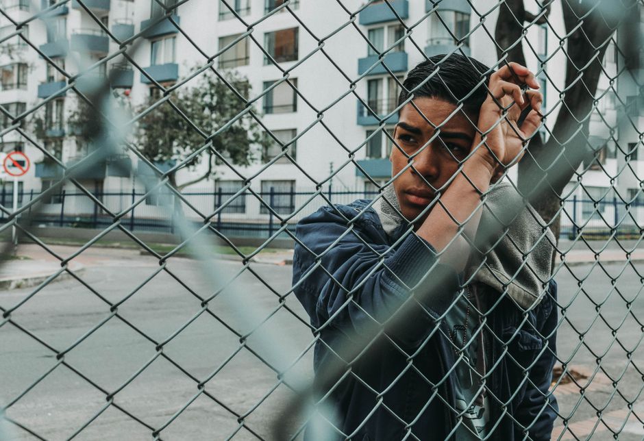 Joven pensativo se encuentra detrás de una cerca de eslabones en un entorno urbano, evocando un sentido de contemplación.
