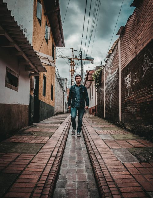 Free Young man in denim jacket walking down a narrow brick alley in Bogota, Colombia. Urban fashion scene. Stock Photo