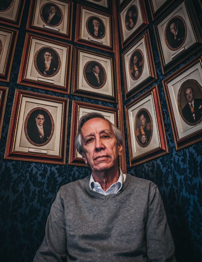 An older man seated in a gallery of framed portraits — photo by Jose Vasquez on Pexels