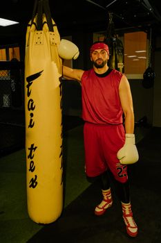 Male boxer in red sportswear and gloves poses beside a punching bag in a gym.