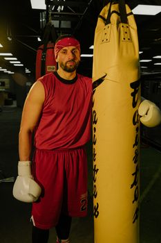 A robust male boxer in red training gear poses with a punching bag in a dimly lit gym setting.