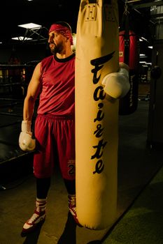 A male boxer in red gear trains with a punching bag inside a gym.