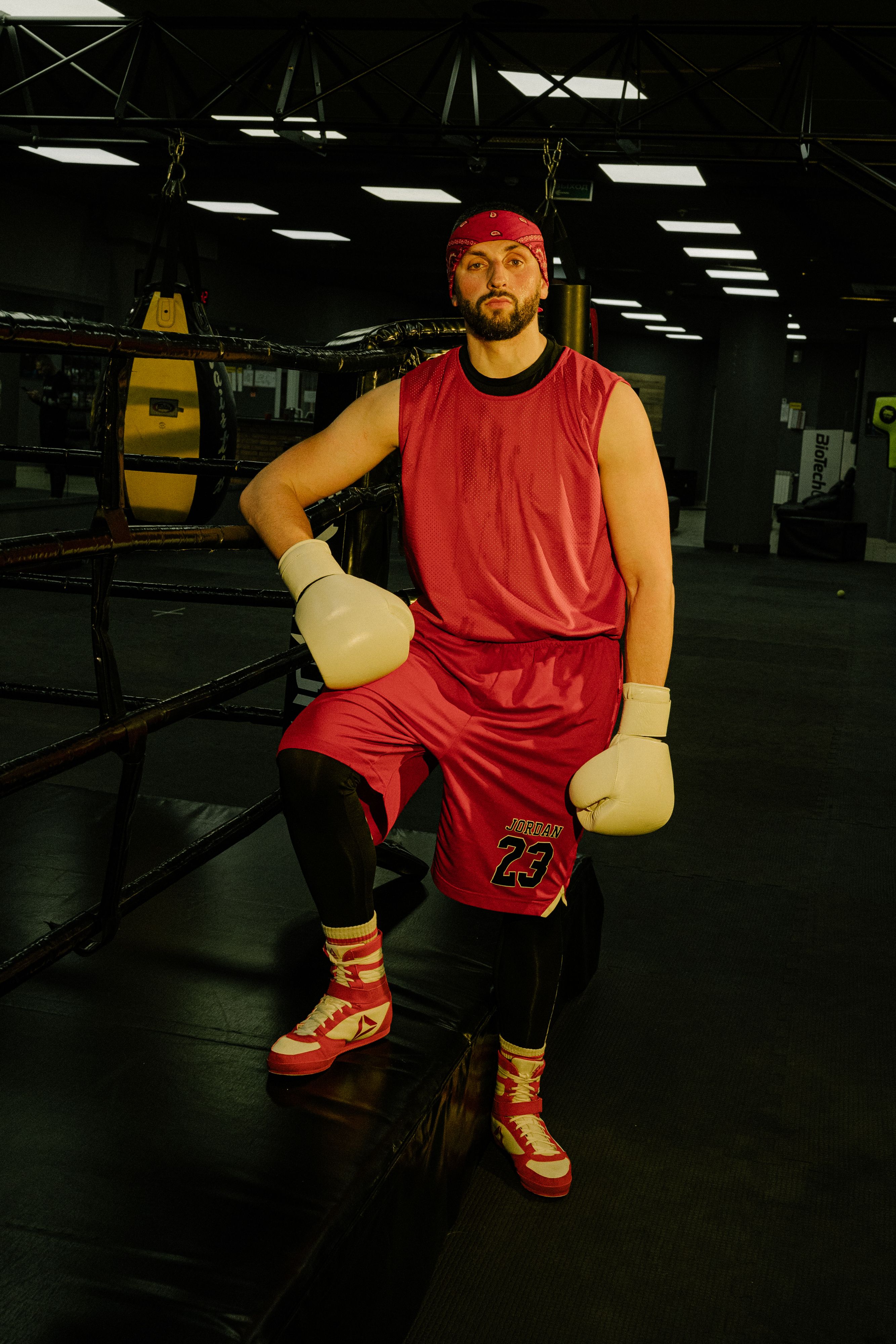 A focused athlete in red gear and boxing gloves prepares in a gym boxing ring.