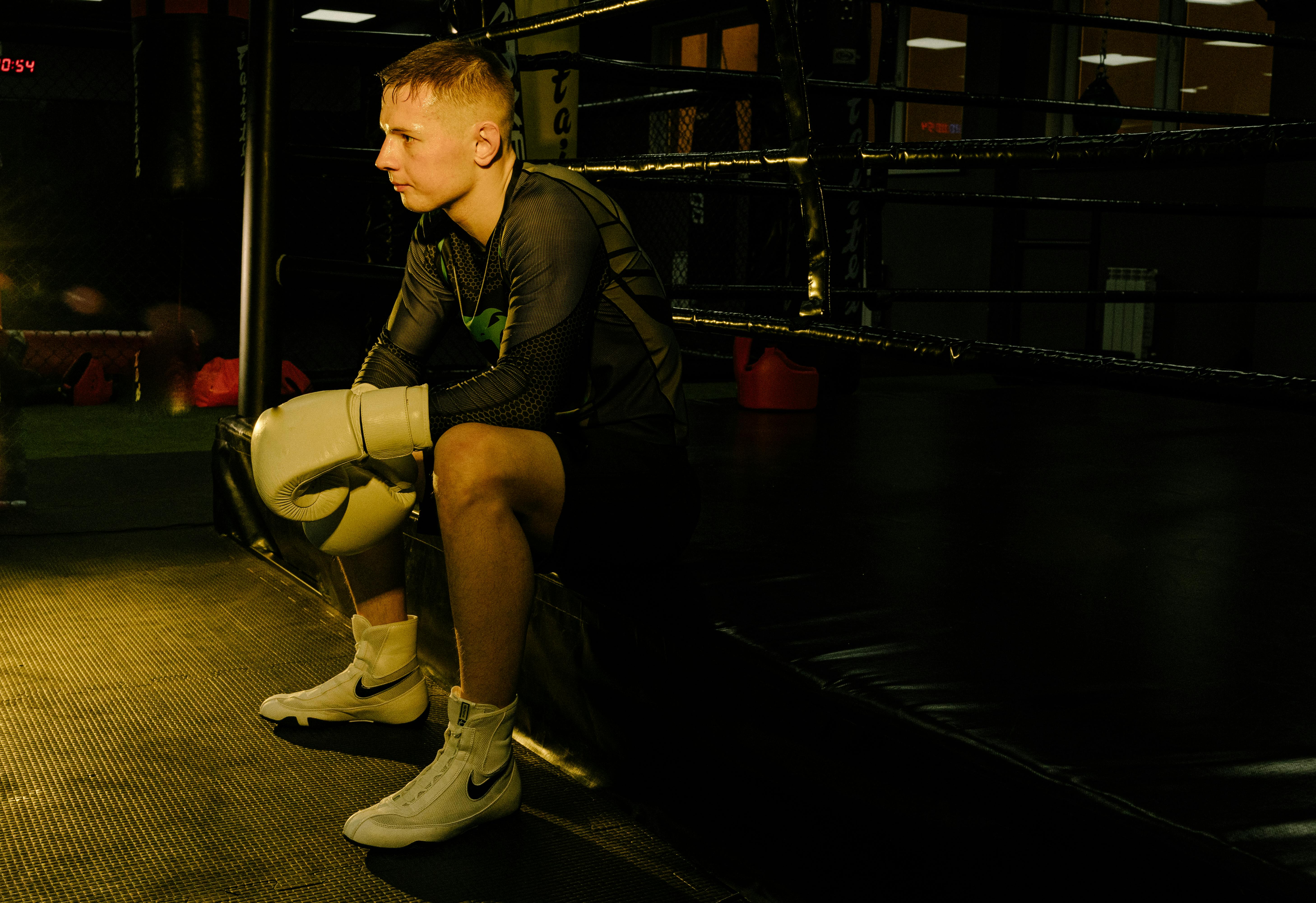 A male boxer sits with gloves on, contemplating in a dimly lit boxing ring.