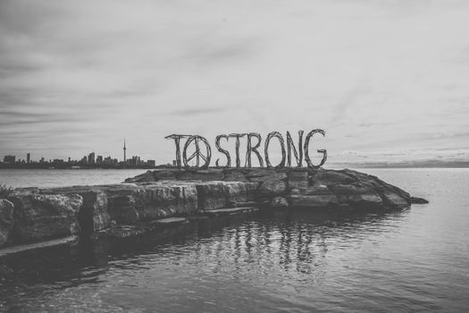 Black and white big inscription To strong with Peace symbol located on stone pier against calm sea and Toronto cityscape