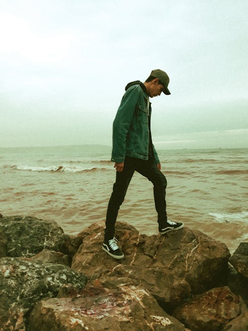 Side view full body ethnic male wearing casual clothes standing on large stones on coast against waving muddy seawater
