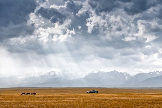 Dramatic view of horses grazing in a vast prairie with distant mountains and a car under cloudy skies.