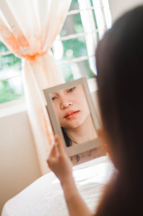 Crop pondering Asian woman reflecting in mirror in flat