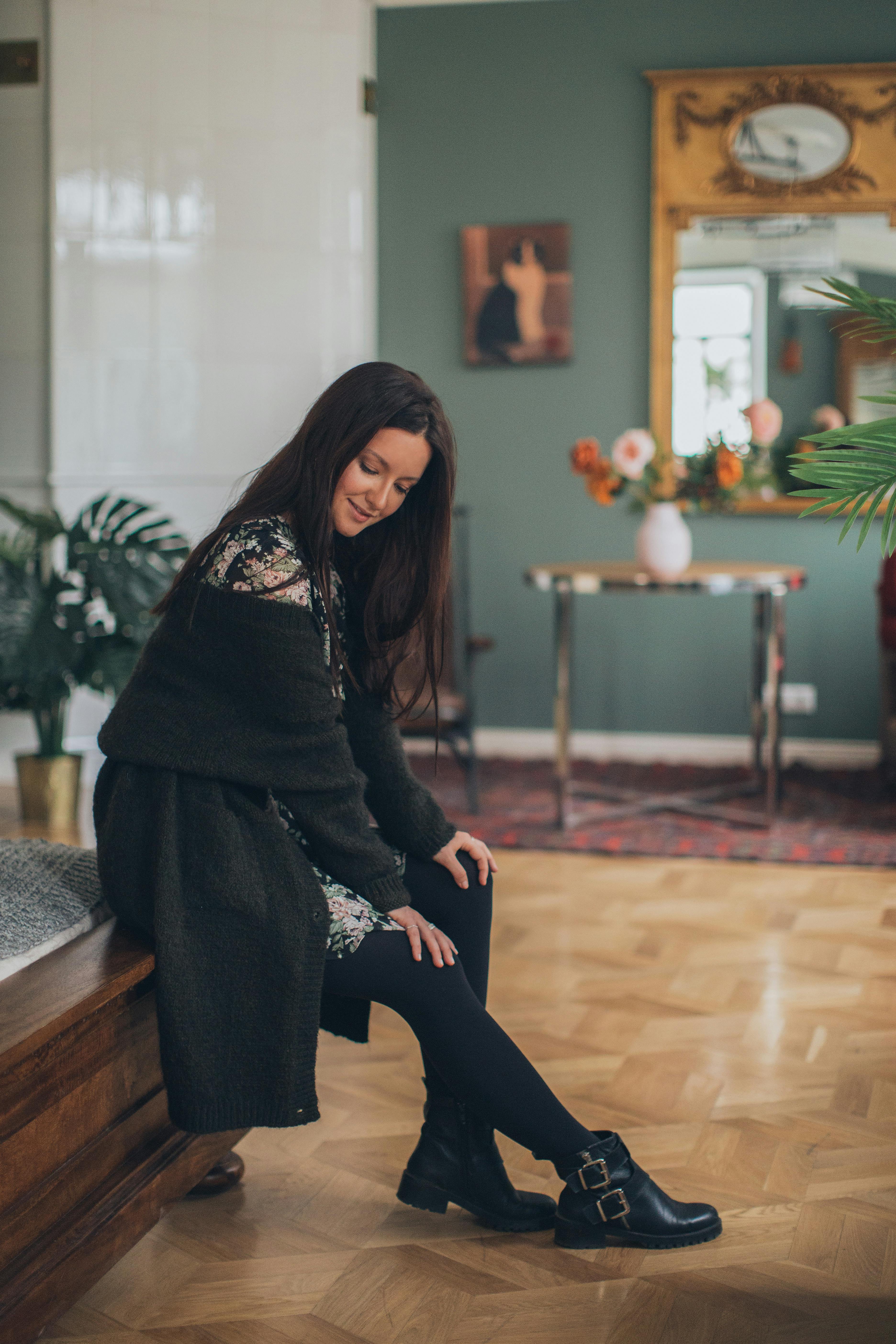 A fashionable woman in a floral dress sits indoors, exuding elegance and style in a modern home interior.