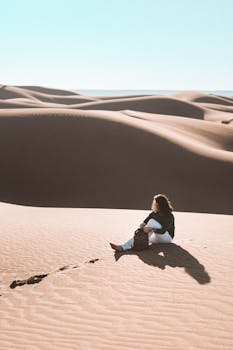 Person sitting on Al Wahat Al Dakhla Desert's sand dunes under the clear sky.