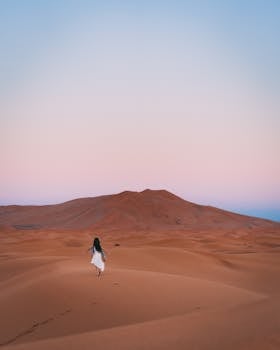 A woman in a white dress walks alone across sand dunes in Al Wahat Al Dakhla Desert during sunset.