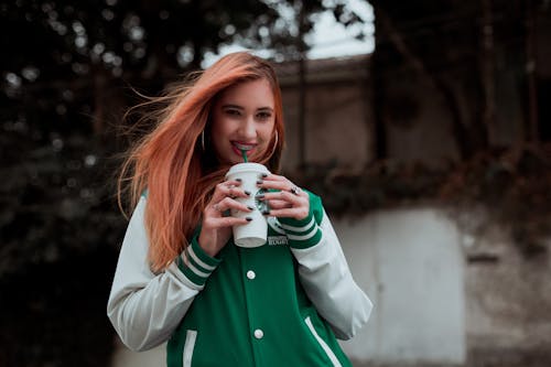 Positive young female drinking hot drink and standing on street while looking at camera