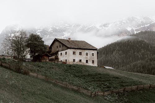 Aged house located on grassy valley near green forest trees growing near mountain ridge under cloudy sky