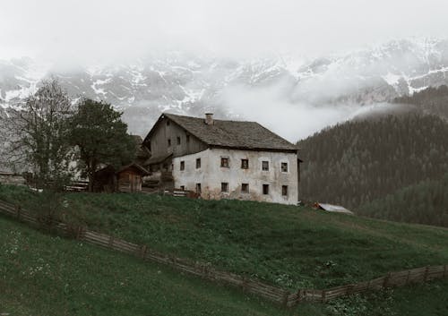 Weathered house located on rough grassy terrain near vast forest trees growing near mountain slope covered with snow in cloudy day