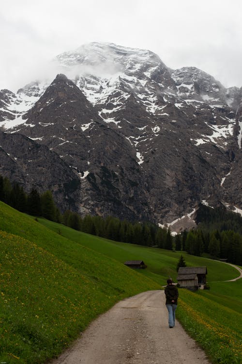 Back view of anonymous traveler walking on narrow path in green hilly valley near powerful snowy mountain ridge and rustic houses