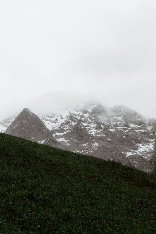 Grey sky and mist above dark green hill and high mountain covered with snow