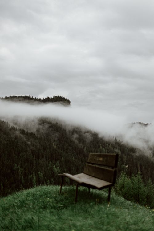 Free Old wooden simple solitude bench on lawn covered with grass next to sky and fog above mountain forest Stock Photo