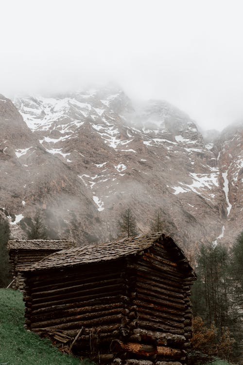 From below of rustic dark brown wooden houses on slope covered with grass by majestic mountain covered with snow under mist