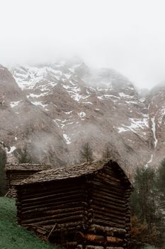 从下方望去，山坡上坐落着一座质朴的深棕色木屋，周围长满了草，而雄伟的山峰则笼罩在薄雾之中，峰顶覆盖着积雪