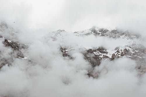 Dense fog covering massive mountain range with snowy rocky slopes on overcast day