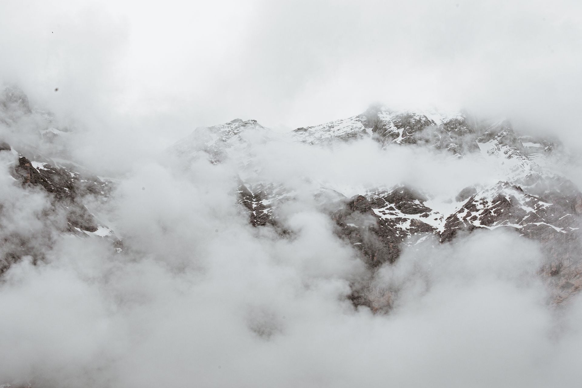 Dense fog covering massive mountain range with snowy rocky slopes on overcast day